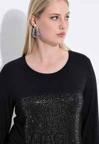 Schwarzes langärmliges Oberteil mit glänzendem, verziertem Textdesign, das mit Strasssteinen "DARLING YOU ARE MAGIC" aufgedruckt ist. Rundhalsausschnitt.