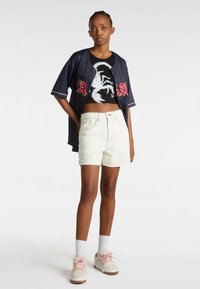 Maillot de baseball marine avec lettres rouges, crop top graphique noir, shorts en denim blancs, et baskets rose clair avec des chaussettes blanches.