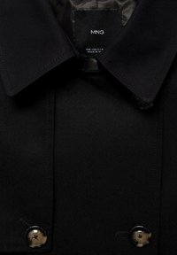 Gros plan sur le col d'un manteau noir avec deux boutons noirs et une étiquette visible indiquant MNG, taille small medium, fabriqué au Vietnam.