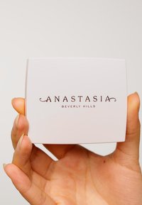 Weißer Kompakt mit dem Text "ANASTASIA BEVERLY HILLS" in Braun. Glatte Oberfläche mit abgerundeten Kanten, gehalten von einer Hand mit natürlichen Nägeln.