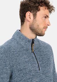Gestrickter Viertelreissverschluß-Pullover in heather-blau mit einer strukturierten Oberfläche, ausgestattet mit einem hohen Kragen und einem kontrastierenden schwarzen Reißverschluss mit einem gelben Zugband.