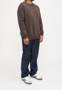 Brązowa bluza z prążkowanymi mankietami i krawędzią, w zestawie z ciemnoniebieskimi jeansami i białymi trampkami z czarnymi paskami. Luźny krój.