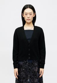 Cardigan preto ajustado com um decote em V profundo, fecho de botões e mangas longas, usado sobre uma saia escura com lantejoulas. Tecido suave com uma textura lisa.