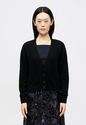 Cardigan noir ajusté avec un décolleté en V profond, fermeture à boutons et manches longues, porté sur une jupe sombre en sequins. Tissu doux avec une texture lisse.