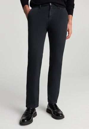 Pantaloni neri con design slim fit, realizzati in tessuto liscio. Presentano tasche laterali e chiusura con bottone, abbinati a scarpe nere lucide.