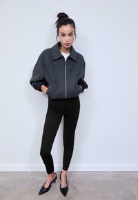 Giacca grigia con zip e colletto strutturato, abbinata a pantaloni neri aderenti e décolleté neri a punta, in piedi davanti a uno sfondo neutro.