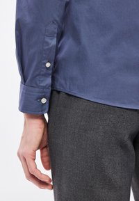 Námornícka modrá košeľa s manžetami na gombíky, detailným prešívaním a hladkou textúrou, nosená so sivými textúrovanými nohavicami. Ruka spočíva na nohe.