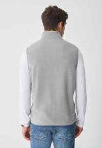 Vesta gri din fleece cu guler înalt, având o textură netedă și fără piese de metal vizibile. Purtată peste o cămașă albă cu mânecă lungă și blugi albastre.