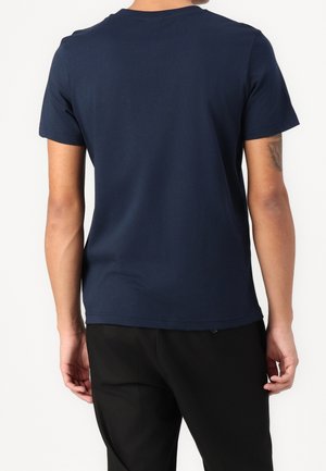 Mand iført en ensfarvet marineblå kortærmet t-shirt og sorte bukser, stående med ryggen mod kameraet foran en hvid baggrund.