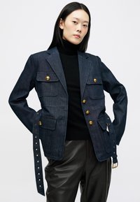 Giacca di jeans blu scuro con bottoni dorati, quattro tasche frontali e una lunga cintura legata con automatici. Sotto, un dolcevita nero.