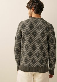 Sweter dzianinowy w stłumionym zielonym kolorze z diamentowym wzorem, teksturowaną powierzchnią, okrągłym dekoltem oraz ściągaczami przy mankietach i u dołu. Noszony z jasnymi spodniami.