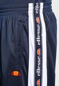 Navy träningsbyxor med vita sidostripes och "ellesse" logotyp, en broderad logotyp i orange och röd, samt sidofickor.