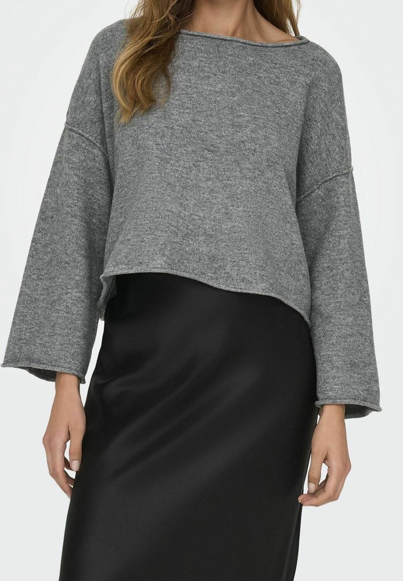 Femme portant un pull gris ample à manches longues et une jupe en satin noire lisse, debout devant un fond clair uni.