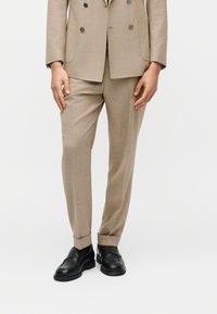 Homme portant un pantalon de costume beige ajusté avec revers et des mocassins en cuir noir, debout devant un fond blanc.