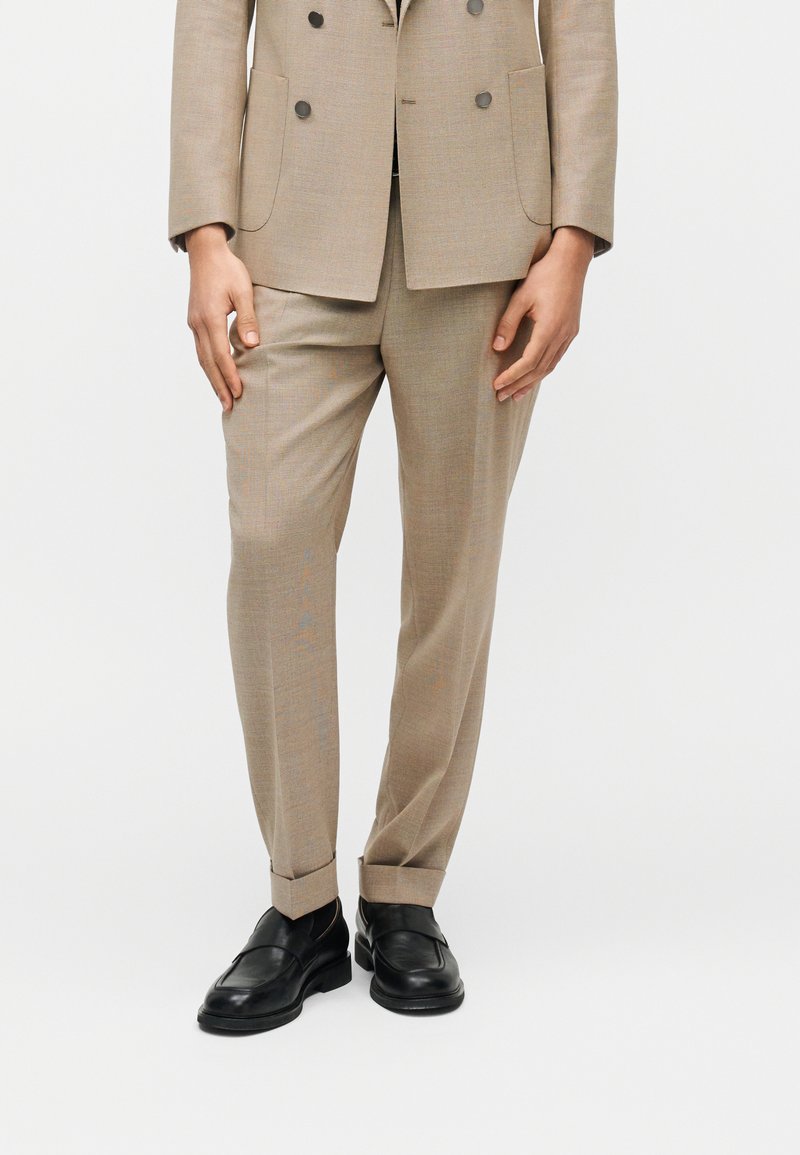 Homme portant un pantalon de costume beige ajusté avec revers et des mocassins en cuir noir, debout devant un fond blanc.