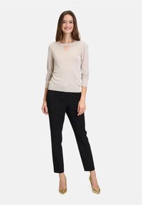 Un pull beige clair avec une découpe en forme de clef, associé à un pantalon noir ajusté et des escarpins dorés, sur fond uni.