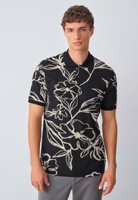 Juodas polo marškinėlis su kreminio atspalvio gėlių raštu, trumpomis rankovėmis ir apykakle. Audinys atrodo lygus ir lengvos tekstūros.