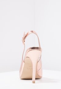 Sandale à talons hauts en rose doré avec une finition métallique brillante, sangle de cheville ajustable et une silhouette élégante et arrondie. Design minimaliste.