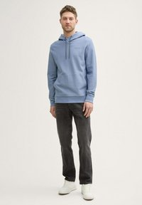 Felpa blu in cotone con cappuccio regolabile, tasca frontale e polsini a coste, abbinata a jeans scuri e sneakers bianche.