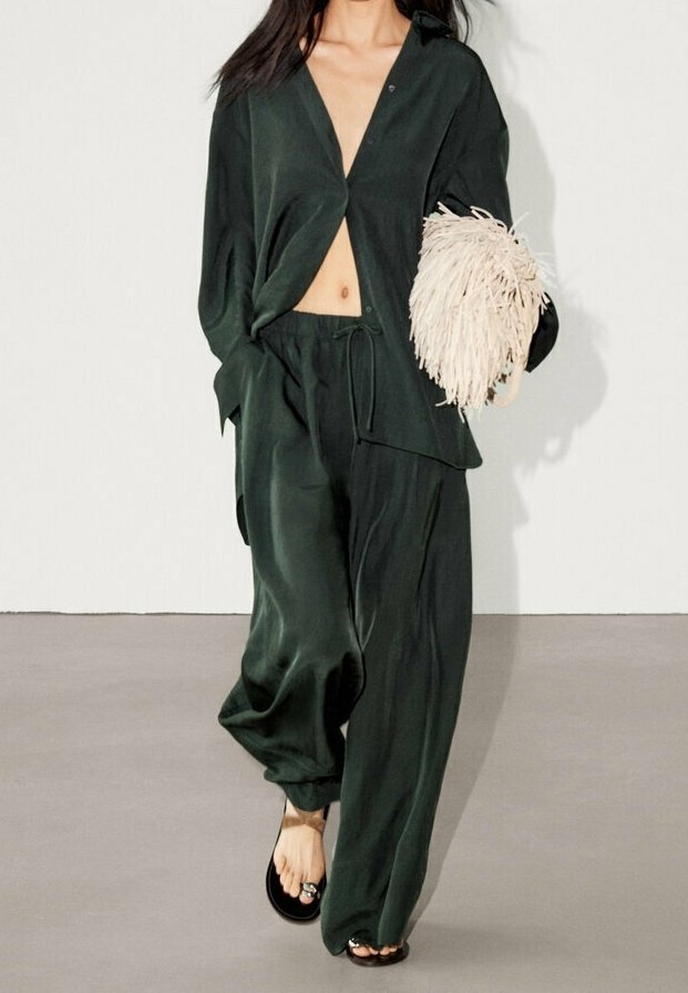 Tummanvihreä satiiniasu, jossa on löysä, napitettava paita ja nyörillä kiristettävät housut. Asustettu teksturoidulla, kermanvärisellä clutch-laukulla.