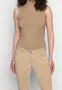Beige žebrovaný bodysuit s vysokým výstřihem, spárovaný se světle béžovými skinny džíny s předními kapsami a stříbrným zapínáním na knoflík.