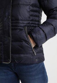 Main dans la poche zippée d'une veste noire matelassée, portée sur un jean bleu, avec des boutons-pression visibles à l'avant et une bordure grise.