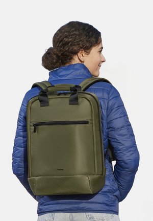 Olive green backpack with a matte finish, featuring a front zip pocket and padded straps, worn over a blue puffer jacket.