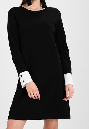 Woman wearing a black long-sleeve dress with white cuffs and black double-button details, standing against a plain background.