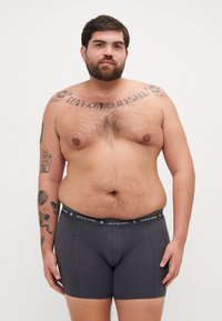 Graue Boxer-Briefs mit einem schwarzen Bund, der "JACK & JONES" trägt. Aus Baumwollmaterial, enganliegendes Design und glatte Textur. Modell mit sichtbaren Tattoos.