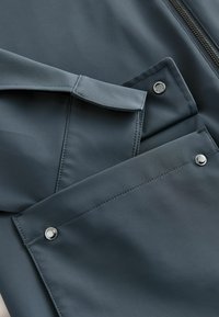 Funkcionāls pelēks auduma žakete ar gludu tekstūru, rāvējslēdzēja aizdari un spiedpogām divās lielās priekšējās kabatās.