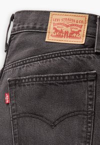 Poche arrière d’un jean Levi's noir avec une étiquette rouge Levi's sur la couture latérale et un patch en cuir beige représentant deux chevaux et un texte.
