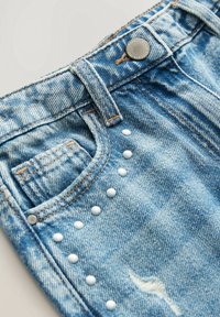 Gros plan sur un jean en denim bleu clair avec une poche avant, un bouton en métal, une passants de ceinture et une rangée de clous ronds blancs près de la poche.