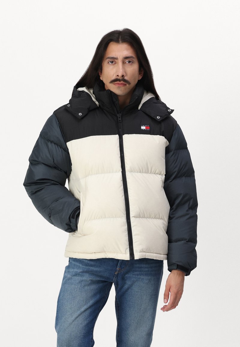 Chaqueta acolchada con un diseño a rayas en negro y crema, que incluye capucha, cremallera frontal y detalle del logo en el pecho. Tejido suave y texturizado.