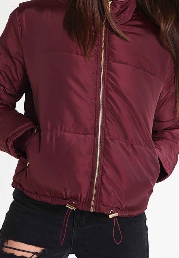 Mujer vestida con una chaqueta de plumas burdeos con cremallera dorada, con las manos en los bolsillos, combinada con jeans negros rotos.