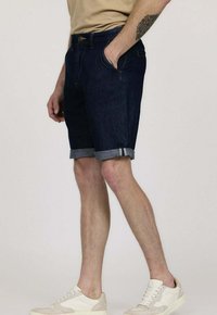Shorts en denim foncé avec un ourlet roulé, présentant des poches avant et une fermeture à bouton. Associés à des baskets de couleur claire.