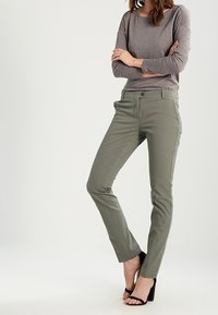 Schmal geschnittene olivgrüne Hose aus elastischem Stoff mit flacher Vorderseite, Gürtelschlaufen und vorderen Taschen. Kombiniert mit einem grauen Langarmshirt.