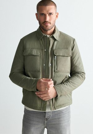 Olive green jacket with a collar, zippered front, and two chest pockets. Made from smooth fabric, features a soft lining and fitted design.