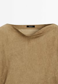 Haut en tissu brun à texture douce avec un col large et un ourlet légèrement irrégulier. Présente une petite étiquette au niveau du col.