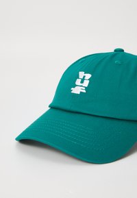 Boné de beisebol turquesa com logotipo branco bordado, aba curva, tecido texturizado e coroa estruturada. Costura reforçada visível.