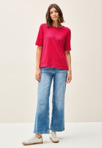 T-shirt rouge à manches courtes avec un graphique circulaire de texte, associé à un jean large bleu clair et des baskets blanches avec des semelles en gomme.