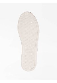Suola di una scarpa con superficie in gomma текстurizzata, colore beige chiaro, forma arrotondata e nome del brand "GUESS" in rilievo.