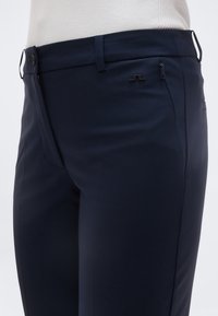 Κοντινό πλάνο σκούρων μπλε παντελονιών με θηλιές για ζώνη, κουμπί και μικρή τσέπη με φερμουάρ στη δεξιά πλευρά, φορεμένα με λευκή ριμπ μπλούζα.