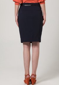 Jupe crayon bleu marine en tissu lisse, avec une fente arrière et une silhouette slim, associée à des sandales à talons hauts orange.