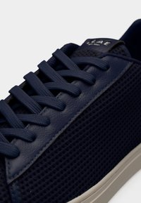 Σκούρο μπλε αθλητικό παπούτσι με υφασμάτινο πλέγμα, χοντρές κορδόνια στο ίδιο χρώμα, δερμάτινη πλευρά με οπές για κορδόνια και λευκή καουτσούκ σόλα, εμφανιζόμενο σε κοντινή γωνία.