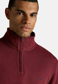 Bordeaux kwart-rits pullover in zachte stof. Heeft een hoge kraag en subtiele ribbelstructuur. Klein blauw logo op de borst.