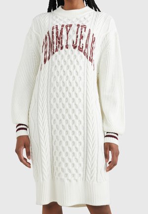 Persona que lleva un vestido de suéter de punto tipo cable blanco con letras burdeos "Tommy Jeans" y puños a rayas en las mangas largas.