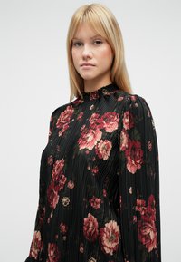 Svart plissért bluse med rysjekrave, med store røde og rosa blomsterprint. Lange ermer med lett puffdetalj.