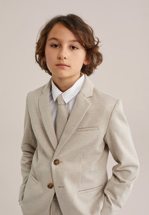 Noor inimene keskmise pikkusega pruunide juustega, kandes beežist ülikonda, valget särki ja sobivat lipsu, seisab ühevärvilise tausta ees.