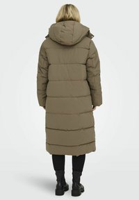 Longue doudoune olive verte avec un design matelassé, capuche, manches avec bord-côte et poches latérales. Portée sur un pantalon noir et des bottes.
