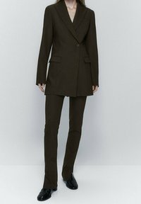 Blazer marron sur mesure avec un bouton unique, des revers crantés et deux poches devant, associé à un pantalon slim assorti et des chaussures noires.